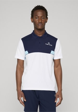 Mand iført marineblå og hvid Sergio Tacchini polo-shirt og marineblå shorts, stående mod en ensartet lys grå baggrund.