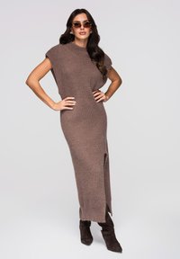 Robe midi en maille marron avec manches courtes, texture côtelée, fente latérale, associée à des bottines en daim marron foncé et des lunettes de soleil.
