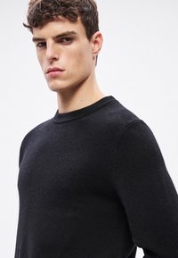 Μαύρο πλεκτό πουλόβερ με στρογγυλή λαιμόκοψη και υφή υφάσματος, εφαρμοστή σιλουέτα και ελάχιστες ραφές. Λείο, απαλό εμφάνιση.