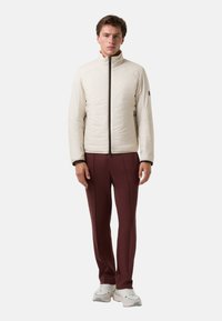 Helle beige Steppjacke mit hohem Kragen und Reißverschluss vorne, kombiniert mit dunkelroten Slim-Fit-Hosen und weißen Sneakers.