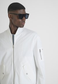 Chaqueta bomber blanca con cuello alzado, dos bolsillos frontales y un bolsillo en el brazo con cremallera. Presenta una textura suave y acentos metálicos.