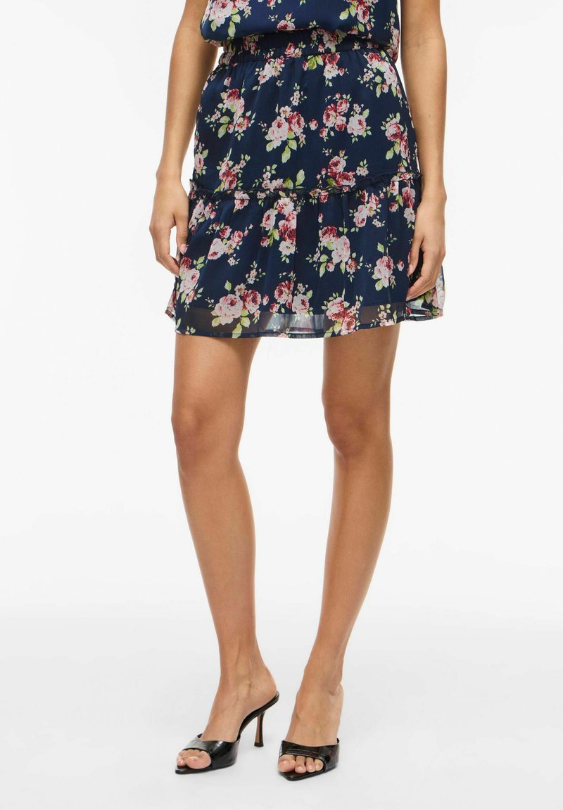 Femme portant une mini-jupe fleurie bleu marine à volants et des sandales à talons hauts noires ouvertes à l'avant, debout devant un fond blanc uni.
