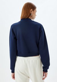 Sudadera corta de marina con mangas largas, puños y dobladillo de canalé, textura suave, combinada con pantalones de corte relajado de color claro.