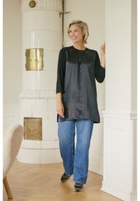 Tunique en satin noir à manches trois-quarts, associée à un jean large bleu et des chaussures à imprimé léopard ; debout contre un mur blanc.