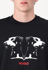 Black T-shirt featuring a white graphic of two dog profiles. The word "HUGO" is printed in red at the bottom. Soft cotton fabric.