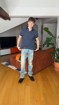 Χαμογελαστός νεαρός άνδρας με ριγέ πόλο και ανοιχτόχρωμο τζιν στέκεται στο σαλόνι με καφέ καναπέ, μεγάλο πράσινο φυτό και τηλεόραση στο φόντο.