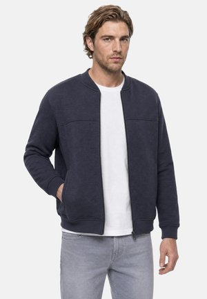 Homme aux cheveux châtain clair portant une veste zippée bleu foncé, un t-shirt blanc et un jean gris clair, debout devant un fond uni.