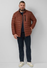 Bruine pufferjas met een ritszak, geborduurde textuur en hoge kraag. Draag het over een marineblauw shirt, in combinatie met donkere jeans en witte sneakers.