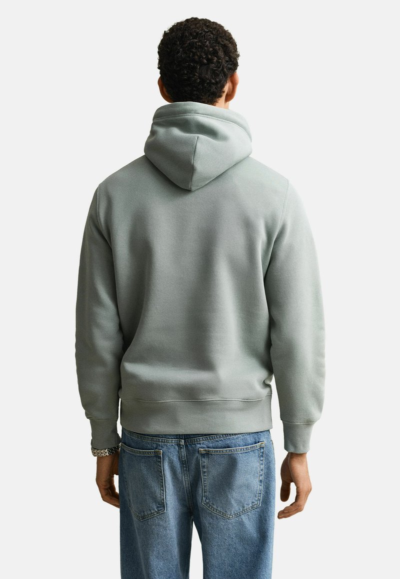 Uomo visto da dietro che indossa una felpa con cappuccio grigio chiaro e jeans blu, in piedi contro uno sfondo chiaro uniforme.