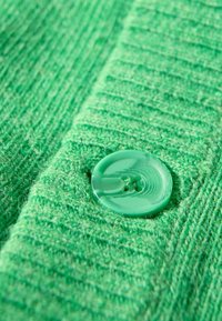 Groene gebreide stof met een gestructureerd ribpatroon, voorzien van een grote groene knop met een spiraalontwerp, gedetailleerde stiksels zichtbaar.