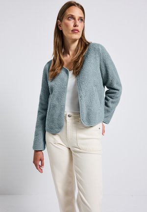 Veste en polaire texturée bleu clair avec un design court, devant ouvert et manches longues, associée à un pantalon beige taille haute.