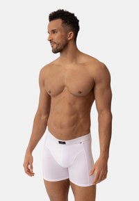 Bílé bavlněné boxerky s kontrastním šedým pasem, se štítkem s logem. Hladký povrch a klasický střih. Model pózuje bokem.