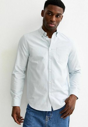 Mężczyzna w jasnoniebieskiej koszuli na guziki, pod którą ma białą podkoszulkę, w niebieskich dżinsach, stoi na tle białej ściany, patrząc w prawo.