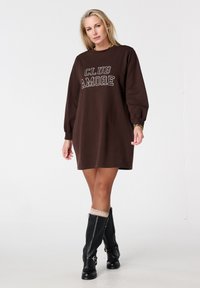 Bruine sweatshirtjurk met de tekst "CLUB AMORE" in het wit. Heeft lange mouwen en een losse pasvorm. Draagt bijpassende zwarte kniehoge laarzen.