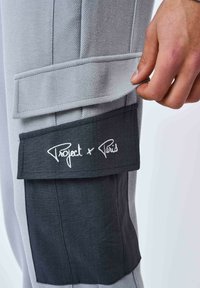 Pantaloni cargo grigi con tessuto testurizzato, dotati di una tasca laterale nera etichettata "Project x Paris" con cuciture bianche.