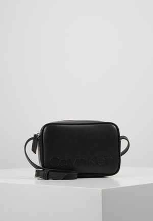 Sac bandoulière rectangulaire noir Calvin Klein avec logo embossé à l'avant et sangle ajustable, présenté sur une surface blanche.