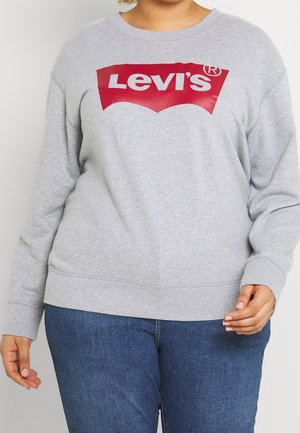 Persoon draagt een grijze Levi's trui met een rood-wit logo, gecombineerd met een blauwe spijkerbroek, staand tegen een effen achtergrond.