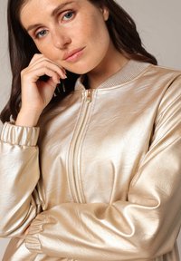 Veste bomber en métal doré avec col et poignets côtelés, présentant une texture lisse et brillante ainsi qu'une fermeture éclair.