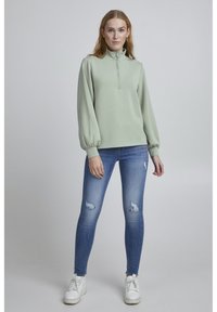 Hele roheline, pika varrukaga pullover kõrge kaelusega ja poolvoodriga, koos siniste kulunud kitsaste teksade ja valgete tennistega.