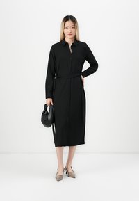 Black long-sleeve button-up dress with a belt, made of soft fabric; paired with a black handbag and beige block-heeled shoes.