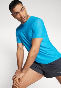 Camisola atlética leve azul com mangas curtas, decote redondo, textura suave e detalhe do logótipo na frente, combinada com calções pretos.