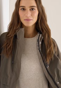 Jeune femme aux longs cheveux bruns portant un pull beige et une veste vert olive non zippée avec des cordons noirs.