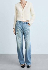 Cream-colored cropped cardigan with textured fabric and three gold buttons, paired with light blue distressed straight-leg jeans. Black heel shoes.