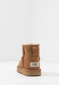 Une botte UGG en suède marron unique avec une doublure en laine visible, vue de dos sur une surface blanche avec un fond blanc.