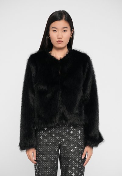Veste en fausse fourrure noire avec un col arrondi, des manches longues et une coupe courte. Portée sur un pantalon noir scintillant.