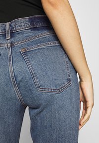 Persona que lleva puestos unos jeans de mezclilla azules con el bolsillo trasero visible y parte de una camiseta negra, con el brazo relajado a su lado.