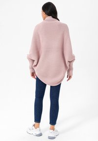 Übergroßer gestrickter Pullover in Hellrosa mit geripptem Muster, ausgestattet mit weiten Ärmeln und einem geschwungenen Saum, kombiniert mit dunkelblauen Skinny-Jeans und weißen Sneakers.