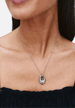 Ciondolo ovale astratto, liscio e argento, su una catena sottile indossato intorno al collo di una donna con una maglia nera con scollo a spalle scoperte.