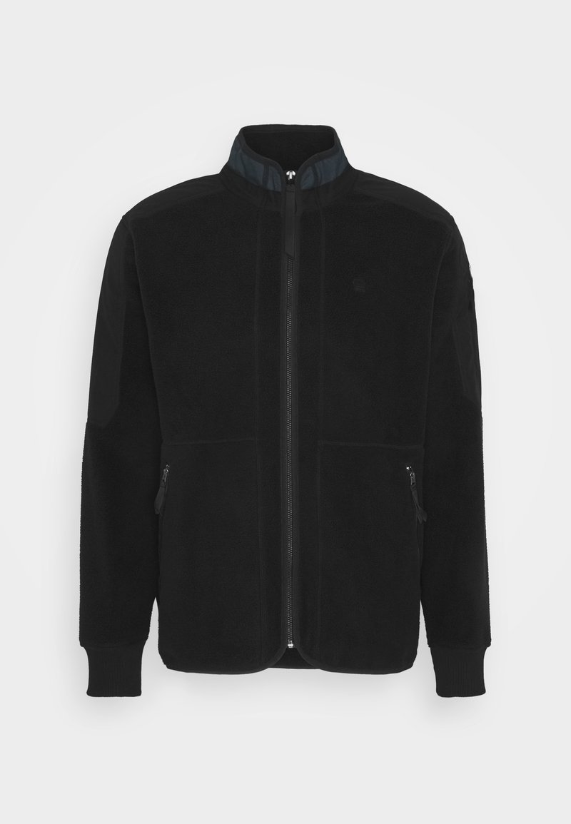 Giacca in pile nera con colletto alto, zip frontale e due tasche laterali con zip. Presenta un tessuto testurizzato nero a contrasto sulle maniche.