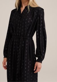 Zwarte jurk met een kraag met knoopsluiting, lange pofmouwen en een stoffen riem. Heeft een gestructureerd patroon en een subtiele glans.