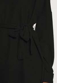 Čierne pletené šaty s viazaným opaskom v páse, dlhými rukávmi a voľným strihom. Textúra sa zdá byť mäkká a hladká, s jednoduchým designom.