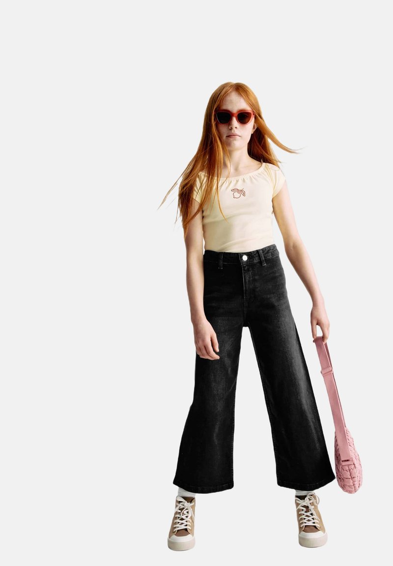 Lichtgele getailleerde T-shirt met bloemenborduursel, zwarte wijde jeans, beige sneakers en een roze schoudertas. Rode zonnebril maakt de look af.