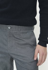 Férfi szürke öltönynadrágban és fekete kötött pulóverben, derékrész látható, jobb keze a combján pihen, fehér háttér előtt.