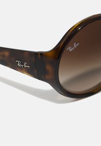 Óculos de sol tortoise castanho com um design robusto, lentes castanhas em gradiente e logótipo prateado de lado, apresentando uma textura plástica suave.