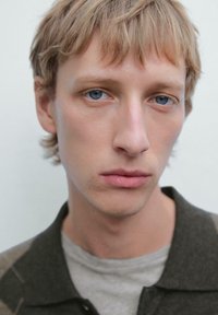 Jong persoon met kort blond haar en blauwe ogen, gekleed in een grijs shirt en een donker geblokte jas, tegen een lichte achtergrond.