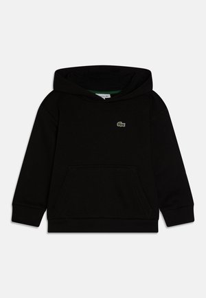 Sudadera negra con bolsillo tipo canguro, logo verde de Lacoste, pantalones negros, chaqueta bomber verde y zapatillas negras con suelas blancas. Gorra azul.