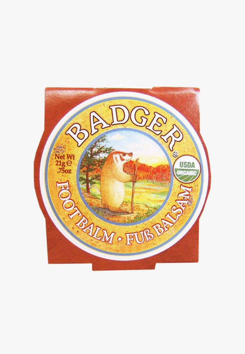 Boîte ronde ornée d'une étiquette colorée avec une illustration de blaireau. Fond rouge, accents dorés, texte indiquant "Baume pour les pieds de blaireau, USDA Organic."