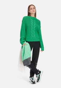 Grüner Strickpullover mit Zopfmuster, lockerer Passform und langen Ärmeln, kombiniert mit schwarzen Hosen und einem mehrfarbigen Schal. Athletische Schuhe runden den Look ab.