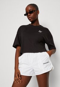 Maglietta nera cropped con logo Puma, abbinata a pantaloni corti bianchi con il testo "puma sports". Tessuto in cotone morbido, vestibilità relax e tasche laterali.