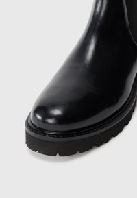 Bottine en cuir noir à bout arrondi, surface texturée et semelle épaisse noire avec un motif de semelle.