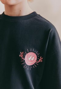 Svart sweatshirt med en rosa smiley-ansikte grafik som har uppsträckta händer och en solstråle-design på vänster bröstområde.