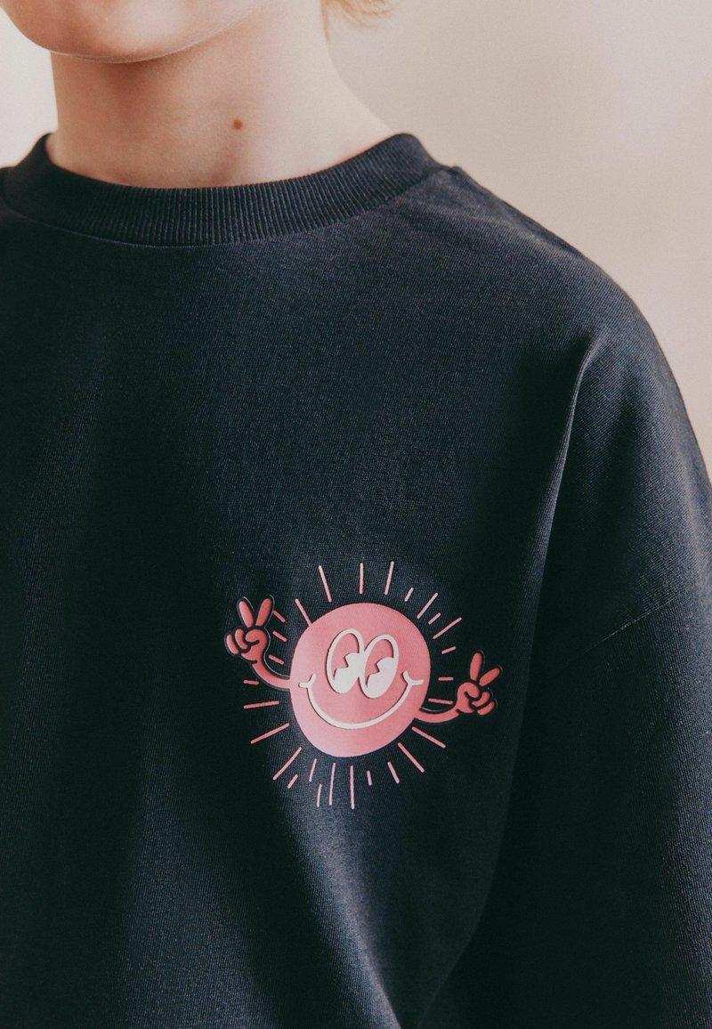 Svart sweatshirt med en rosa smiley-ansikte grafik som har uppsträckta händer och en solstråle-design på vänster bröstområde.