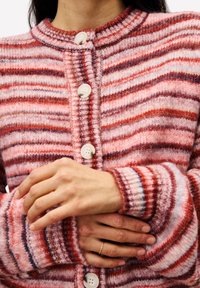 Femme portant un cardigan en maille rayé rose et rouge avec des boutons blancs, les bras croisés à la taille, une main montrant une fine bague en or au doigt.