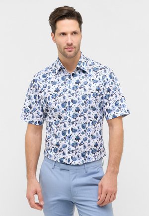 Mand iført en kortærmet hvid skjorte med blå blomsterprint og lyseblå bukser, stående foran en ensfarvet baggrund.