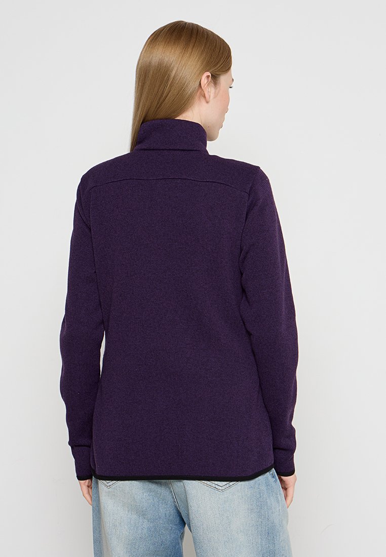 Frau mit glattem, hellbraunem Haar, die eine dunkelpurpurne Fleecejacke und hellblaue Jeans trägt, mit dem Rücken zu einer schlichten weißen Wand.