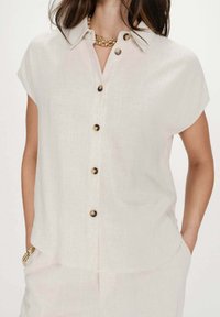 Mujer con camisa beige de manga corta y botones, collar de cadena dorada y varias pulseras doradas, con las manos en los bolsillos de un pantalón beige.